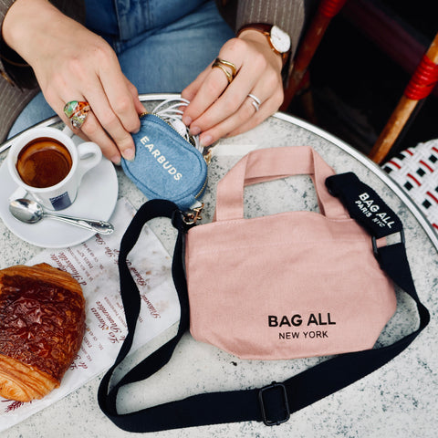 Bag-all Mini Tote Bag in blush pink with black strap, shown on marble table with coffee and croissant, featuring New York branding and compact design for everyday essentials
