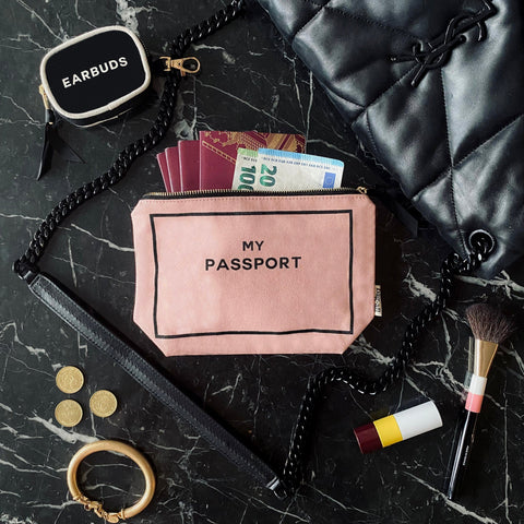 Bag-all Passport Pouch in Pink/Blush shown on black marble surface with travel essentials - passport, currency, earbuds case, and makeup brush, featuring gold zipper and cotton fabric