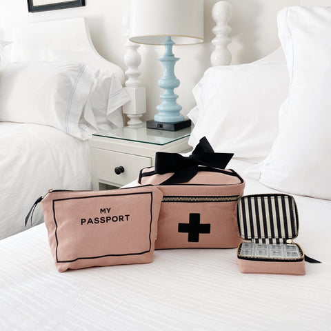 Bag-all First Aid Travel Gift Set 3-Pack featuring pink cotton passport pouch, medical storage box with cross symbol, and striped pill case on white bedding. Perfect for organized, stylish travel.