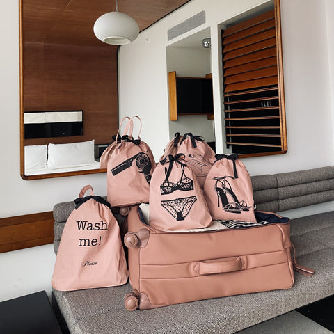 Bag-all Pink Hair Dryer Travel Bag with matching lingerie and laundry organizer bags on grey couch, featuring blush pink cotton material, drawstring closures, and black printed designs with bathroom mirror reflection