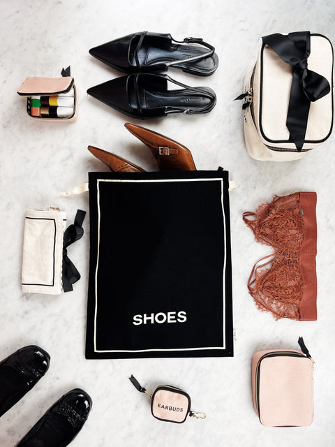 Bag-all Couture Shoe Travel Bag in black with elegant white trim, displayed with luxury accessories including leather shoes and travel essentials on marble surface