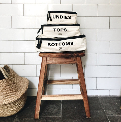 Bag-all Cotton Packing Cubes, Print, 3-pack Cream: Three organized travel cubes labeled Undies, Tops, and Bottoms stacked on wooden stool, perfect for efficient packing and clothing organization