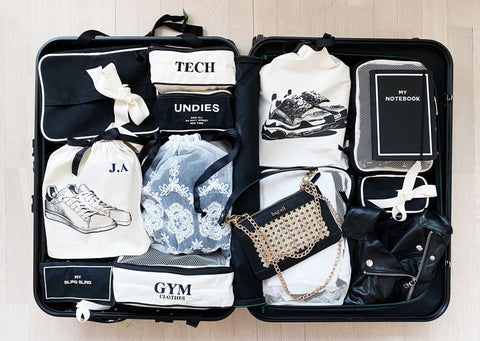 Bag-all Cotton Packing Cubes 3-pack Cream - Organized suitcase interior showing labeled storage bags for clothes, tech, gym gear, and accessories in black and white color scheme