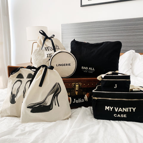 Bag-all Black Pumps Shoe Bag with stylish high heel print, shown among travel organizers and vanity cases on white bedding, featuring cream canvas material and black drawstring closure