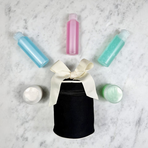 Black round carry on liquids case with refillable bottles and cream jars on marble surface, tied with a white ribbon bow.