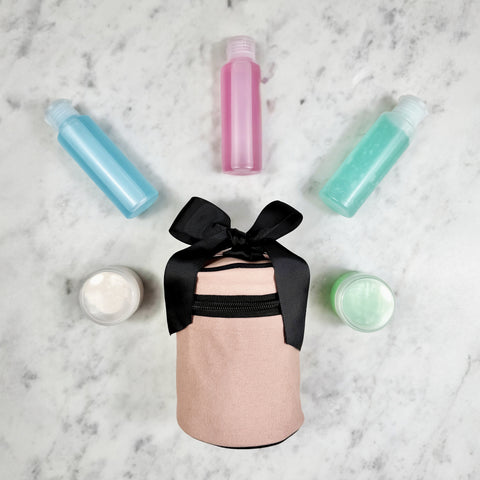 Pink blush round carry on liquids case with black bow, surrounded by refillable travel bottles and cream jars on marble