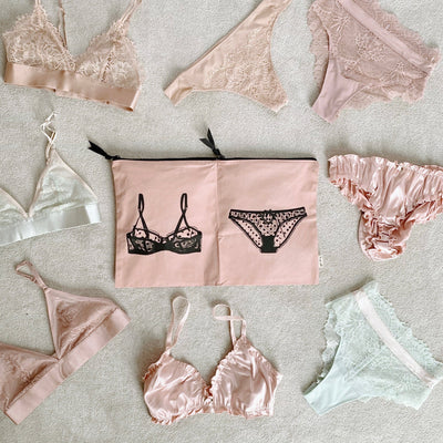 Bag-all Double Lingerie Packing Pouch in pink/blush features black lingerie illustrations, surrounded by delicate lace bras and panties in soft pink and white tones, perfect for organized travel storage