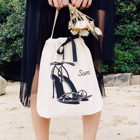 Bag-all High Heel Sandal Shoe Bag in cream cotton with black stiletto illustration and navy ribbon, shown against black dress with dried flowers, perfect for shoe storage and travel organization