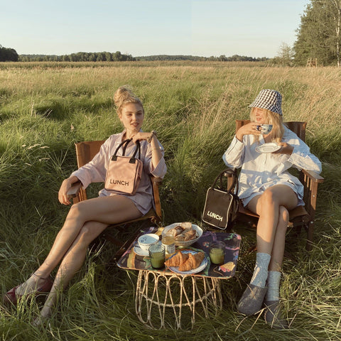 Bag-all Black Insulated Lunch Box shown in outdoor picnic setting with vintage-style wire table, featuring packed sandwiches and drinks, displaying practical portability and stylish design in natural meadow scenery