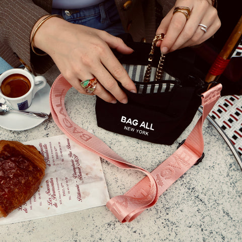 Bag-all Mini Tote Bag with Strap in black cotton, featuring interior pocket, shown with coffee and pink accessories on marble surface. Sustainable everyday carry solution from New York.