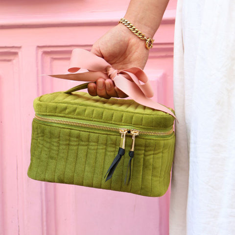 Hand holding a personalized quilted green velvet makeup cosmetic box with a pink ribbon handle against a pink background