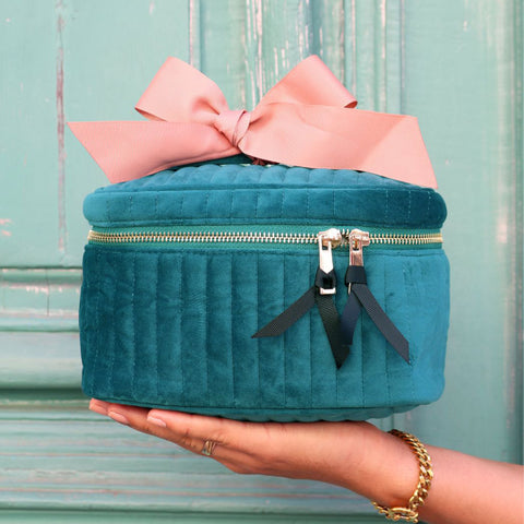 Quilted teal velvet makeup cosmetic box with gold zipper and pink ribbon held in hand against turquoise background