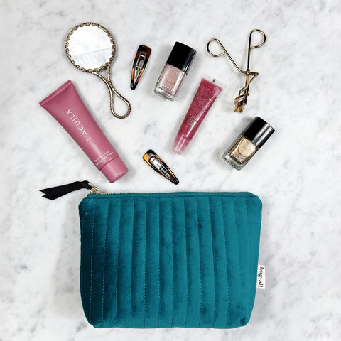 Teal quilted velvet makeup pouch with coated lining surrounded by beauty products and hair clips on marble surface