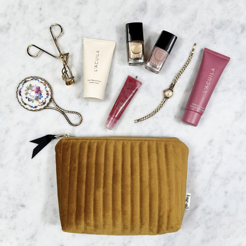Quilted yellow velvet makeup pouch with zipper, surrounded by nail polish, cream tubes, lip gloss, eyelash curler, and watch on marble surface