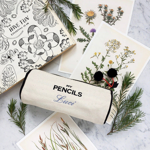 Bag-all cream canvas pencil case with 'My Pencils' text and custom name embroidery, displayed with botanical illustrations and pine sprigs on marble surface