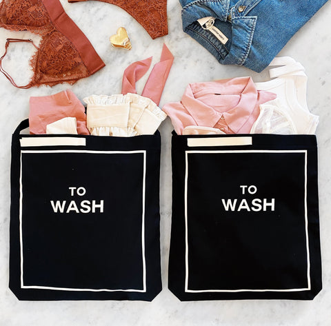 Bag-all To Wash Laundry Bag in black cotton with white border detail and text, shown filled with pink and neutral clothing items on marble surface, perfect for organizing dirty laundry