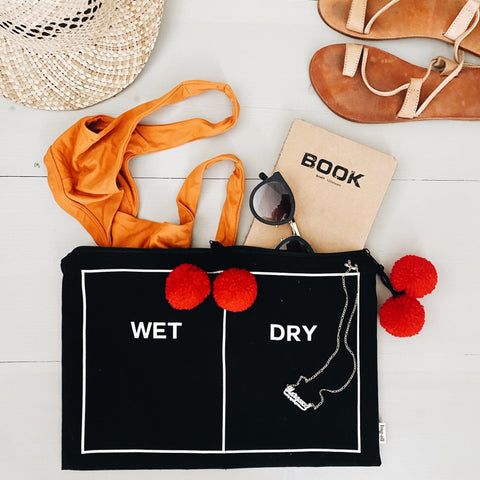 Bag-all Wet/Dry Pouch Black with red pom-poms - dual compartment beach and pool organizer bag featuring white text labels and decorative trim, styled with summer accessories