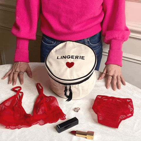 Bag-all Round Lingerie Case in cream canvas with black trim and heart detail, shown with red lace lingerie pieces and cosmetics on white surface