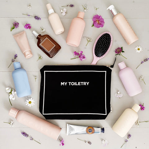 Bag-all Toiletry Pouch with Coated Lining in black, surrounded by beauty products, hairbrush and delicate flowers on light wooden surface. Waterproof travel organizer for cosmetics and toiletries.