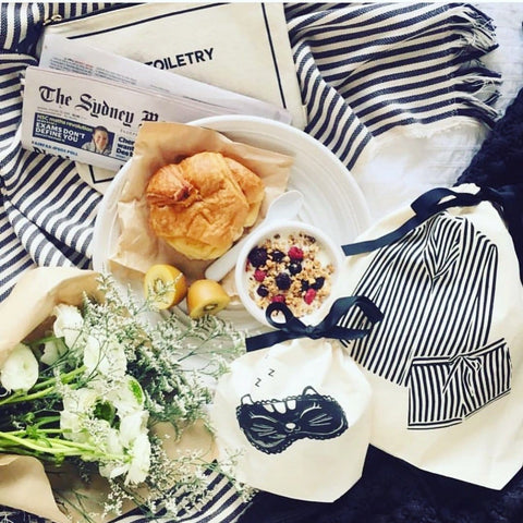 Bag-all Striped Pajamas Travel Bag in cream, styled with breakfast, fresh flowers, and newspaper, showcasing classic navy and white stripes against white bedding, perfect for travel organization